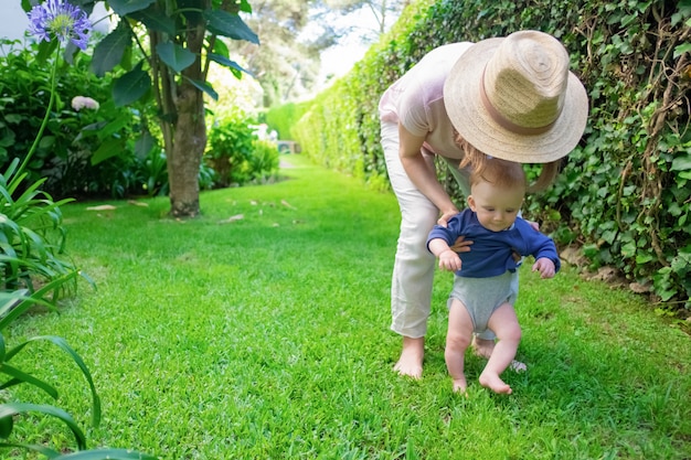 Bambino sveglio in camicia blu che fa i primi passi con l'aiuto della mamma e sorridente. Giovane madre in cappello che tiene neonato sull'erba. Primi passi a piedi nudi