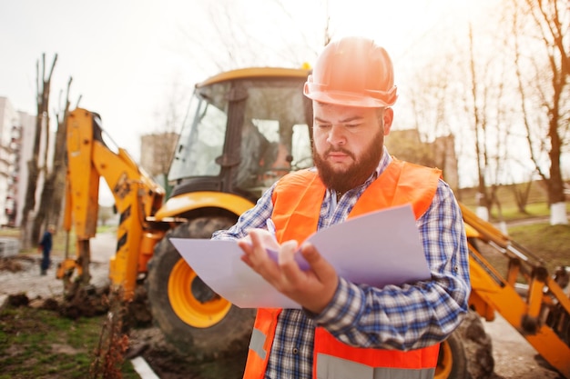 Barba brutale lavoratore uomo vestito operaio edile in casco arancione di sicurezza contro traktor con carta piano a portata di mano