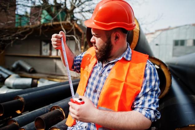 Barba brutale lavoratore uomo vestito operaio edile in casco arancione di sicurezza vicino a tubi d'acciaio con metro a nastro a portata di mano