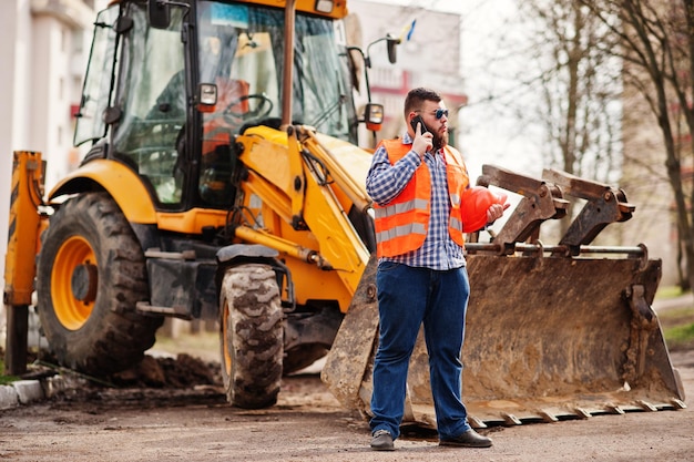 Barba lavoratore uomo vestito operaio edile in occhiali da sole casco arancio di sicurezza contro il trattore con il telefono cellulare a portata di mano