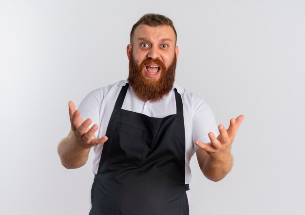 Barbiere barbuto professionista uomo in grembiule confuso e frustrato con le braccia alzate in piedi sopra il muro bianco