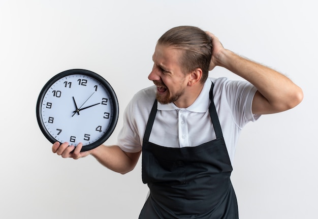 Barbiere bello giovane arrabbiato che indossa la tenuta uniforme e guardando l'orologio con la mano sulla testa isolata su bianco