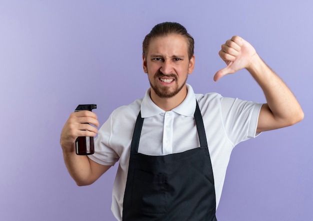 Barbiere bello giovane dispiaciuto che indossa la bottiglia spray tenuta uniforme e che mostra il pollice verso il basso isolato su sfondo viola