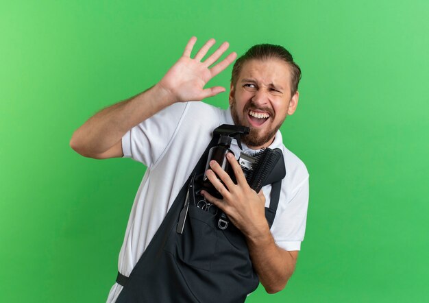 Barbiere bello giovane infastidito che indossa pettini di tenuta uniformi, flacone spray, tagliacapelli guardando a lato e non facendo alcun gesto isolato su verde con spazio di copia