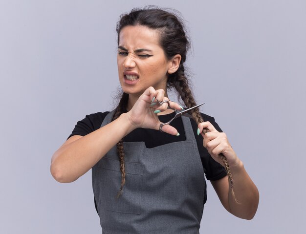 Barbiere femminile giovane arrabbiato in capelli di taglio uniforme con le forbici isolate sulla parete bianca