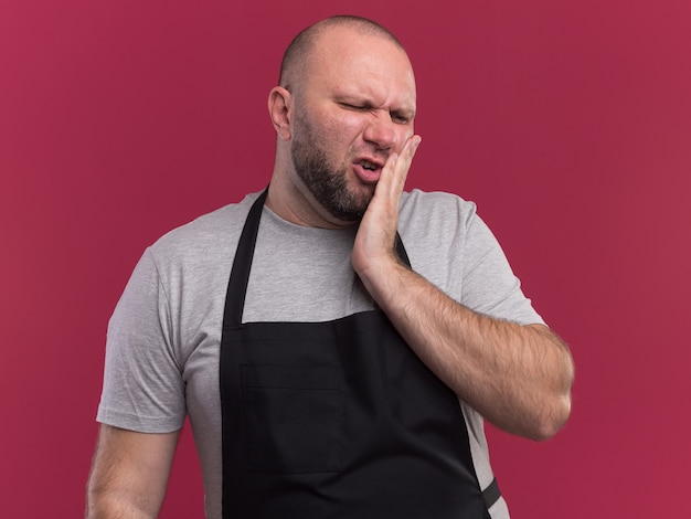 Barbiere maschio di mezza età slavo dispiaciuto in uniforme che mette la mano sul dente dolorante isolato sulla parete rosa