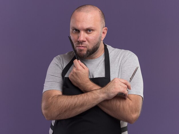 Barbiere maschio di mezza età slavo rigoroso in uniforme che pettina la barba e che tiene le forbici isolate sulla parete viola