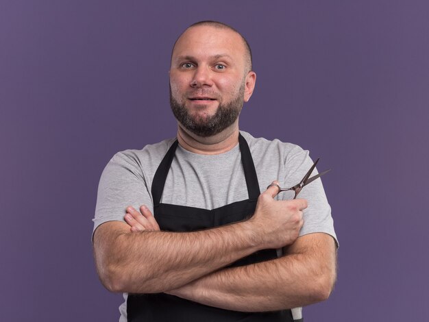 Barbiere maschio di mezza età slavo sorridente in uniforme che tiene le forbici che attraversano le mani isolate sulla parete viola