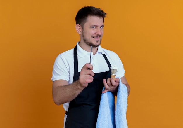 Barbiere uomo in grembiule con asciugamano sulla sua mano tenendo il pennello da barba con schiuma e rasoio guardando la telecamera sorridente fiducioso in piedi su sfondo arancione