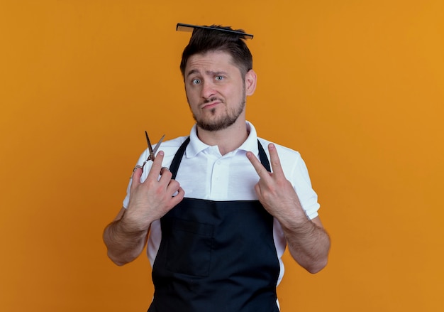 Barbiere uomo in grembiule con spazzola per capelli tra i capelli tenendo le forbici che mostra il numero due guardando la telecamera scontento in piedi su sfondo arancione