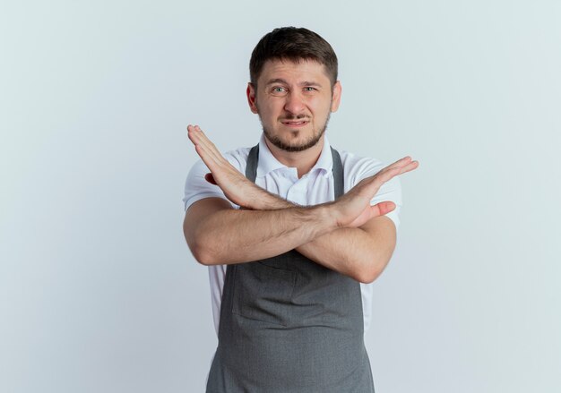 barbiere uomo in grembiule dispiaciuto che mostra il segnale di stop attraversando le mani in piedi sul muro bianco