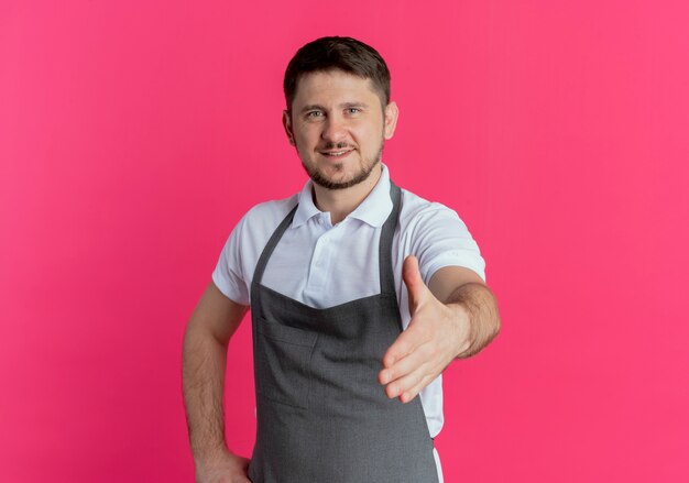 Barbiere uomo in grembiule guardando la telecamera offrendo mano saluto sorridente amichevole in piedi su sfondo rosa