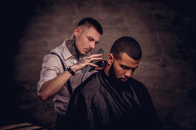 Barbuto afroamericano hipster che si fa tagliare i capelli da un parrucchiere professionista tatuato vecchio stile fa il taglio di capelli.