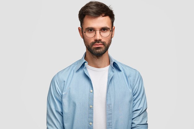 Barbuto giovane maschio sicuro di sé con aspetto piacevole, vestito con camicia blu, guarda direttamente, isolato sopra il muro bianco Libero professionista bell'uomo pensa al lavoro al coperto.