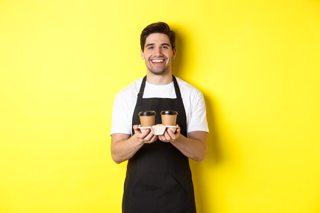 Barista amichevole in grembiule nero che dà ordine da asporto, con in mano due tazze di caffè e sorridente, in piedi su sfondo giallo.