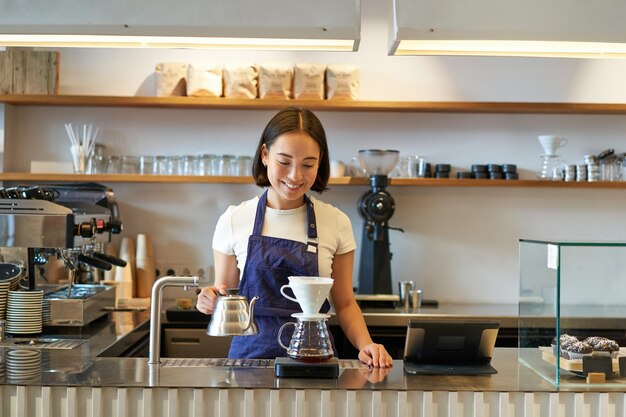 Barista asiatico sorridente della ragazza che lavora nel caffè che prepara il caffè v che prepara l'ordine dietro il bancone