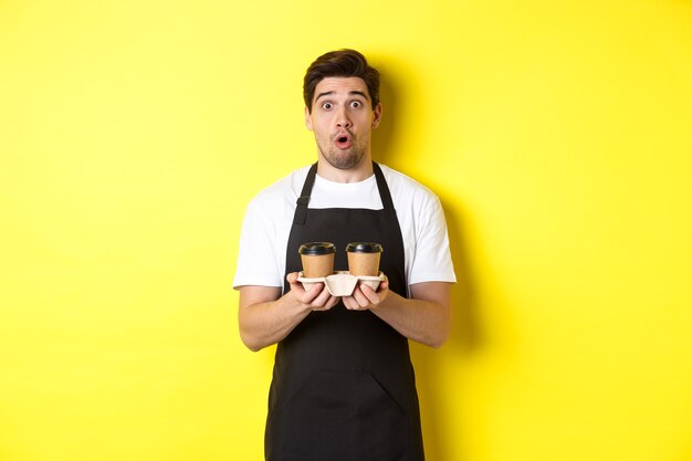 Barista che serve caffè, guardando sorpreso dalla fotocamera, indossa un grembiule nero, in piedi su sfondo giallo.