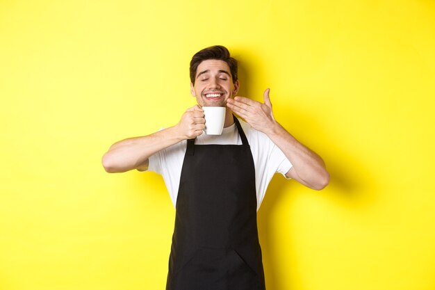 Barista che si gode l'odore del caffè in tazza, in piedi soddisfatto con gli occhi chiusi, indossa un grembiule nero.