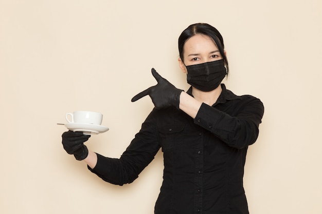 barista femmina in pantaloni camicia nera con caffè marrone essiccato ingredienti tè attrezzature in maschera sterile nera sul muro bianco