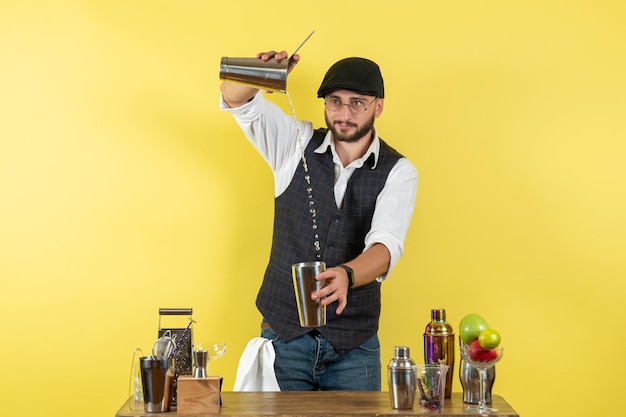 Barista maschio vista frontale davanti al tavolo con agitatori che fanno un drink sul muro giallo bar alcol notte club drink giovanile