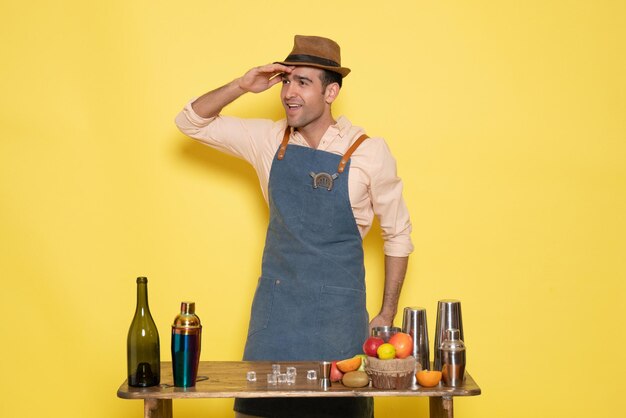 Barista maschio vista frontale davanti al tavolo con bevande e shaker sulla parete gialla beve alcol bar notturno color club male