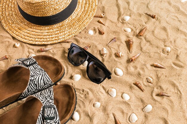 Beachwear estivo, infradito, cappello, occhiali da sole e conchiglie sulla spiaggia di sabbia.