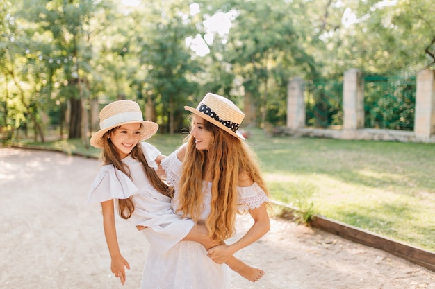 Beata donna con lunghi capelli biondi che trasportano ragazza a piedi nudi in barca alla moda con la natura. Ritratto all'aperto di allegra giovane mamma trascorrere del tempo con il bambino in una giornata estiva.