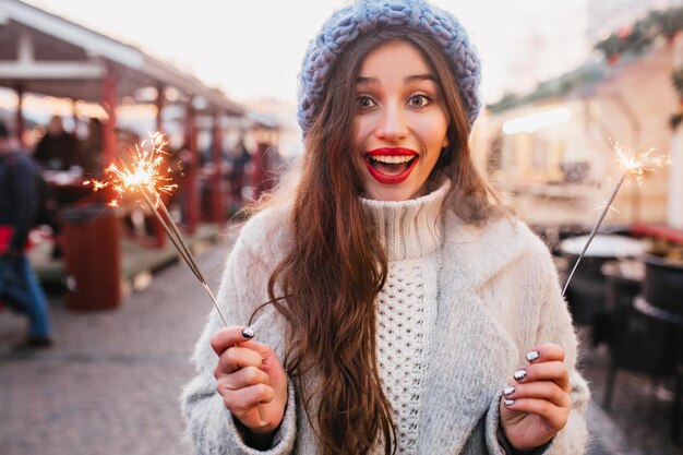 Beata donna dai capelli castani con un sorriso sincero che si gode le vacanze di Natale e posa con lo sparkler. Affascinante ragazza in morbido cappello blu che tiene la luce del Bengala sulla strada.