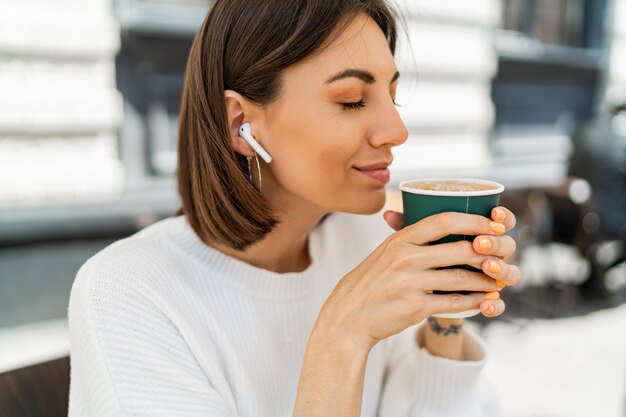 Beata donna dai capelli corti che si gode il cappuccino al bar, indossando un maglione bianco accogliente e ascoltando la musica preferita con gli auricolari