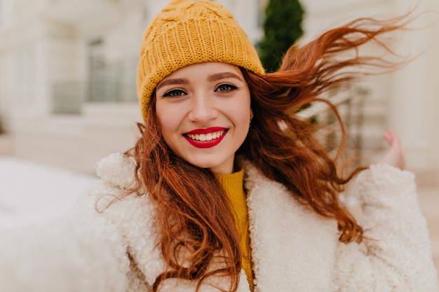 Beata donna dai capelli lunghi con labbra rosse che fa selfie nel fine settimana invernale. Ragazza allegra dello zenzero in cappello che esprime felicità.