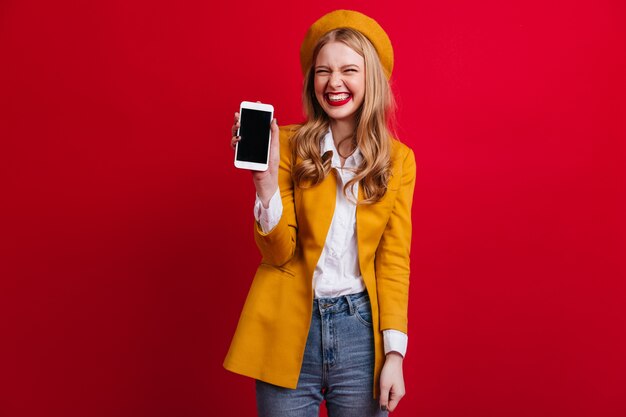 Beata donna francese che tiene smartphone con schermo vuoto. Vista frontale della ragazza bionda elegante in berretto isolato sulla parete rossa.