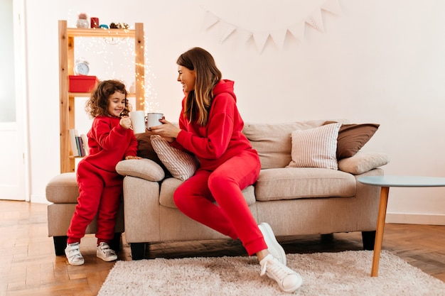 Beata donna in abito rosso, bere il tè con la piccola figlia. Tiro al coperto di sorridente madre e bambino in posa al divano.