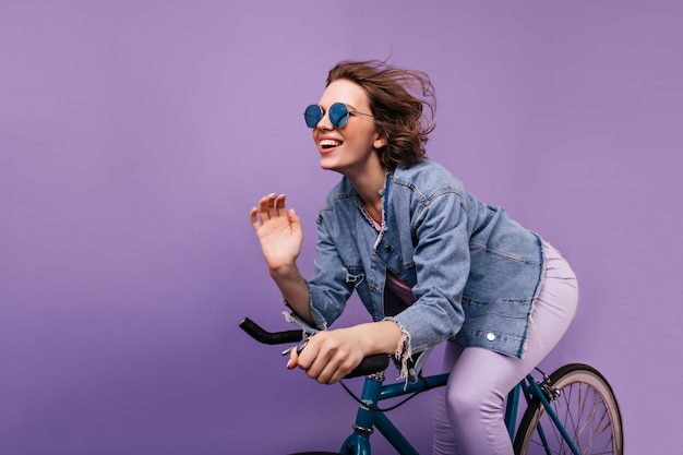 Beata donna in giacca casual in posa sulla bici. Emotiva ragazza dai capelli corti in bicchieri scintillanti in sella alla bicicletta.