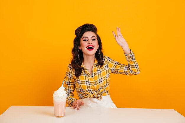 Beata giovane donna con milkshake ridendo su sfondo giallo. Studio shot di positivo pinup girl in brillante camicia a scacchi.