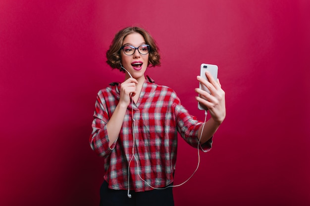 Beata giovane modello femminile indossa occhiali e camicia a scacchi facendo selfie sul muro bordeaux. felice ragazza dai capelli corti con il telefono divertendosi nel tempo libero.