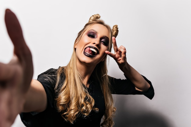 Beata ragazza bionda con labbra nere che fanno selfie in halloween. Incredibile giovane strega divertente in posa sul muro bianco.