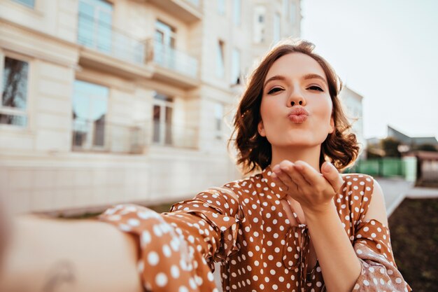 Beata ragazza in abbigliamento vintage che fa selfie sulla strada. Splendida signora caucasica in abito marrone che invia un bacio d'aria