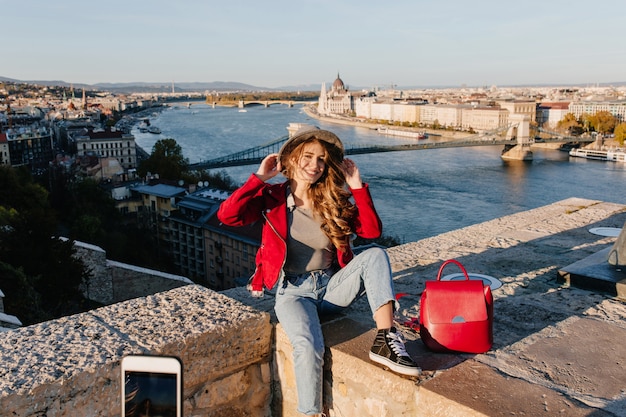 Beata ragazza in abiti casual che si siede sul tetto e tocca il suo cappello