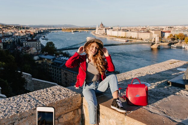 Beata ragazza in abiti casual che si siede sul tetto e tocca il suo cappello