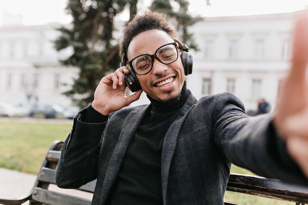 Beato uomo africano dai capelli neri che fa selfie sulla città. Foto all'aperto del ragazzo mulatto che ride felice con l'acconciatura riccia.