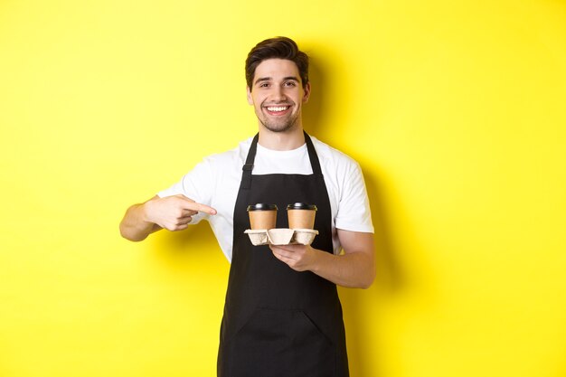 Bel barista che tiene due tazze di caffè da asporto, puntando il dito verso le bevande e sorridendo, in piedi in grembiule nero su sfondo giallo.