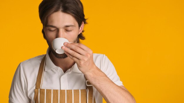 Bel barista in grembiule che beve caffè chiudendo gli occhi con piacere Giovane uomo barbuto che si gode un buon caffè su sfondo colorato