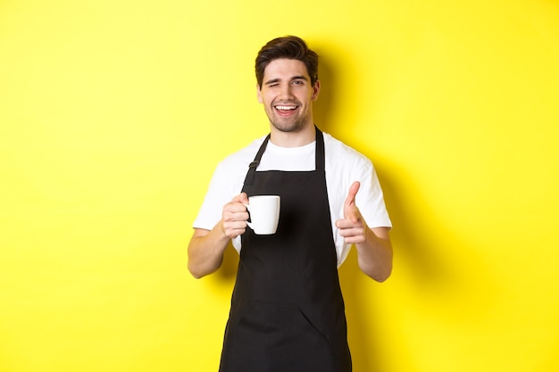 Bel barista in grembiule nero che tiene una tazza di caffè, puntando il dito contro di te, invitando il caffè a visitare, in piedi su sfondo giallo