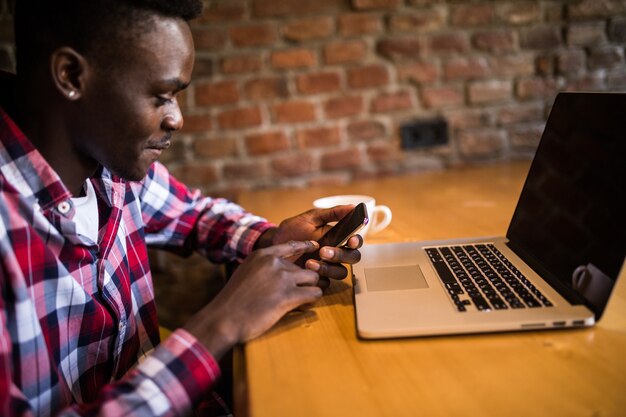 Bel giovane afroamericano sorridente felicemente durante la lettura di sms su smart phone, messaggistica utilizzando la connessione wi-fi gratuita, un caffè al bar