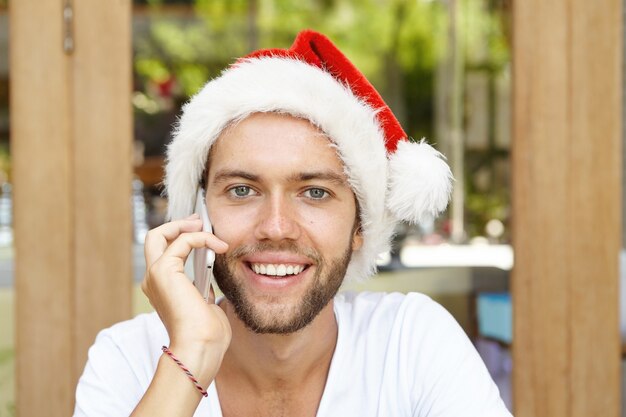 Bel giovane barbuto hipster che indossa il cappello rosso con pelliccia bianca in posa all'interno come Babbo Natale, avendo una bella conversazione telefonica