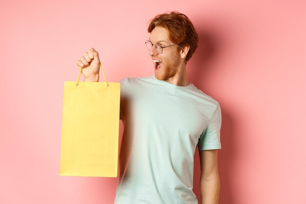 Bel giovane che compra regali, tiene in mano la borsa della spesa e sembra divertito, in piedi su sfondo rosa