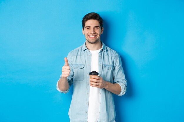 Bel giovane che mostra pollice in su e beve caffè, raccomandando caffè da asporto, in piedi su sfondo blu.