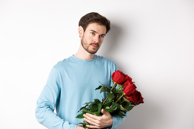 Bel giovane con bellissime rose rosse per il suo amante il giorno di San Valentino, guardando pensieroso, in piedi su sfondo bianco