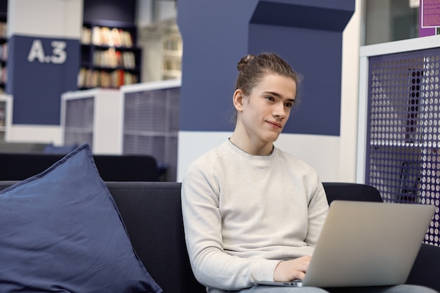 Bel giovane con i capelli alla moda chignon sorridente durante la riproduzione di videogiochi utilizzando wifi ad alta velocità sul computer portatile