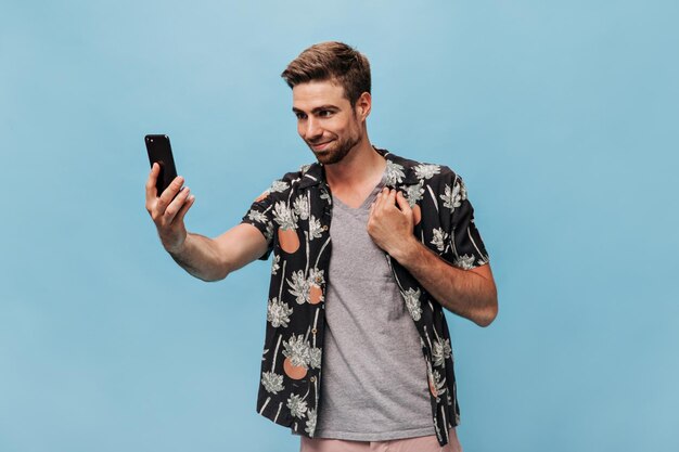 Bel giovane con la barba in abiti eleganti e alla moda che scatta foto sorridendo e posando con lo smartphone su sfondo blu isolato